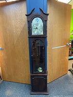 Full height frontal view of wooden grandfather clock standing against wooden double doors. Shows entire clock from bonnet top to base.