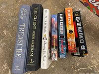 Seven books displayed upright showing titles and authors, a collection from thriller and adventure genres.