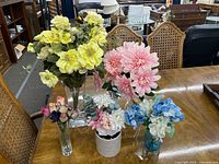 Five artificial flower arrangements on a wooden table in various glass and ceramic vessels.