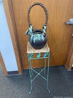 Full view of the ceramic teapot with twisted wicker handle sitting on metal and wicker stand against a wood door.