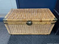 Front view of the rectangular wicker trunk showing the top lid, metal latch, and metal corner protectors.