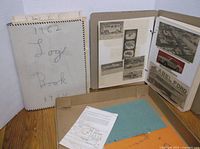 Overview of the scrapbook standing upright with pages visible, showing glued clippings and photos along with its white cover with handwritten title.