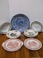 Photo showing all 13 plates and bowls arranged on wooden surface, with the largest blue speckled bowl at the center back and plates with scenic prints at the front.