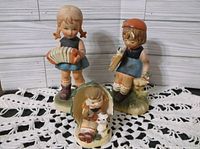 Three porcelain figurines showing children in vintage Hummel-like style arranged on a crocheted lace cloth with wooden plank background.
