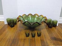 Wide view showing large scalloped green and amber glass bowl with 6 green glasses on sides and 3 shot glasses in front on hardwood floor against white background.