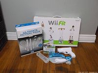 Photo showing boxed Wii Fit Board, Ultimate Action Pack, and game controllers on wooden floor.