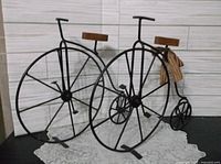 Side view of two vintage-style black metal high-wheel tricycles with wooden seats on a white textured runner and white paneled backdrop.
