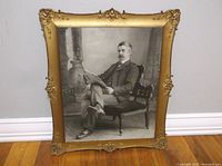 Full view of the gold-colored ornamental plastic picture frame containing the black and white portrait of a seated man holding a newspaper.