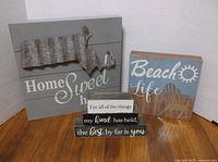 Three wooden decorative signs arranged on a white backdrop and wooden floor: a grey 'Home Sweet Home' sign with metal Massachusetts cutout, a blue 'Beach Life' sign, and a set of stacked blocks with text.
