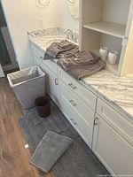 Photo showing brown hand towels and bath towels folded on bathroom countertop next to two clear cups, gray shower rugs on floor, laundry basket, and small brown woven waste basket.