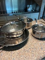 Cookware set displayed on counter showing multiple pots with glass lids and frying pans in polished stainless steel finish.