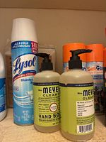 Photo showing Lysol disinfectant spray, two bottles of Mrs. Meyer's lemon verbena hand soap with pump dispensers, and partially visible orange-capped spray cans.