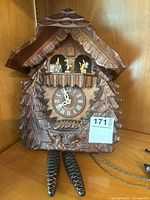 Wooden German cuckoo clock with pine tree carving and pine cone weights on table, front view showing clock face and figurines window.