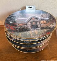 Stack of eight round decorative plates displaying a winter nighttime cottage scene in Thomas Kinkade's painting style.