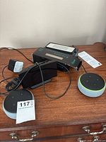 View showing two Amazon Echo Dot smart speakers, a black battery pack or speaker device, and a black alarm clock on a wooden surface.