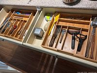 Top view of two kitchen drawers containing assorted kitchen tools including knives, scissors, peelers, spatulas, graters, and other utensils arranged in wooden dividers.