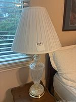 Front view of vintage crystal lamp with white pleated fabric lampshade on wooden bedside table next to bed.