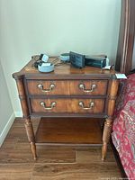 Front view of Ethan Allen wooden nightstand with two drawers, metal handles, turned legs, stained finish, and items placed on top.