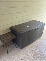 View of the dark brown Suncast outdoor storage box with a small wicker side table beside it.