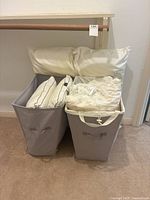 Two large cream-colored pillows, two quilted pillows with brown piping, and two smaller printed pillows inside two gray fabric laundry baskets