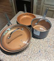 Photo of the three cookware items on a kitchen counter: 10-inch frying pan with lid, 8-inch frying pan without lid, 6-inch pot with lid, all with copper-colored interiors and dark exteriors.