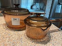 Two amber-colored Pyrex glass pots with lids shown on a kitchen countertop. One pot is smaller (1L) and the other larger (2.5L). Both have long handles and glass knobs on lids.