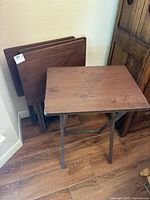 Photo of four foldable wooden TV table trays stacked upright against a wall on wood flooring, showing the rectangular wood table tops and foldable legs.
