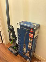Bissell Bolt Lithium Pet upright vacuum and original box standing next to each other on hardwood floor.