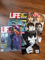 Four LIFE magazines laid out on wooden floor showing covers: Beatles swimming cover, Beatles portraits cover, Paul McCartney at 75 cover, and Live Aid issue cover.