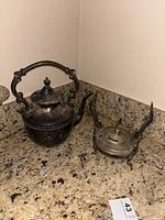 Two-piece ornate silver on copper server set displayed on granite countertop. The set includes a teapot and a chafing dish stand with lid, both showing tarnish and patina.