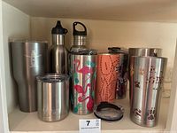 View of all eight sport water bottles and one Yeti stainless steel coffee mug arranged together on shelf, some lids missing.