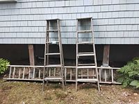 Photo showing two wood step ladders standing upright against a house siding with metal extension ladder laid horizontally on the ground