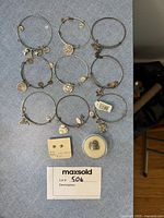 Nine silver-tone Alex and Ani charm bracelets laid out in a grid with varying charms such as crosses, roses, sunflowers, hearts, four-leaf clovers, and symbolic motifs. Also shows two rings: one .925 silver spoon ring and one pearl-accented ring.