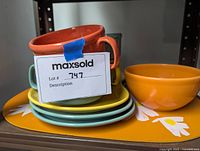 Stack of four cups in orange, yellow, green, and gray stacked on matching colored plates and a yellow floral ceramic tray visible underneath.