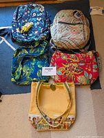 Five assorted quilted fabric bags arranged on carpet, two backpacks and three bags of various styles and colors including Vera Bradley branded items.