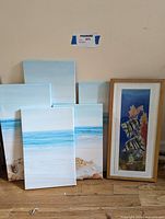 Five wall art pieces including four canvases of beach scenes and one framed painting of fish underwater, all grouped on floor against wall.