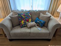 Beige fabric loveseat with four decorative cushions, placed in front of window with beige curtains, showing armrest, cushions, and legs.
