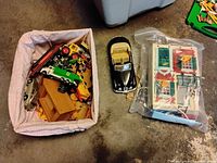 Photo showing a basket with an assortment of small toy vehicles including cars and trucks, a black convertible toy car, and a wrapped small dollhouse or building set toy.