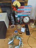 Photo showing a tall clear glass vase with artificial cream and red flowers, a rustic wooden inspirational sign, a round clock with a white face and gold rim, two metallic cone figurines, and part of the floral wreath and photo frame.