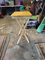 Photo showing one wood folding table with light wood square top and wooden X folding legs, taken in garage setting to depict table condition and style
