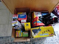 Top view of two cardboard boxes containing assorted boxes of plastic storage bags, sandwich bags, aluminum foil, and plastic wrap, alongside a black plastic bag dispenser.
