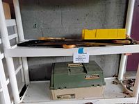 Green and beige Plano fishing tackle box on a shelf with hand saws and tools placed above.