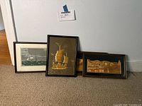 Three framed prints leaning against wall on floor, featuring ships and landscapes.