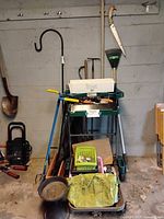 Mixed lot of garden tools including green garden cart, green tote with supplies, and long handled tools leaning against a wall.