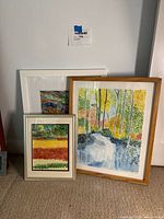 Four framed paintings leaning against a wall: includes a vertical landscape in a light wood frame showing trees and water, a smaller abstract piece with horizontal color bands in a silver metal frame, and other mixed media artworks in white frames. Frames show minor wear.