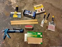 Painting supplies laid out on floor including paint brushes, putty knife, sanding sponge, rulers, caulking gun, steel wool, sandpaper and Velcro sticky back