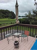 Photo shows beige patio umbrella folded on deck with three small glass-top metal tables and outdoor garden heater with planter pot.
