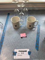 Two white ceramic mugs with black Playboy Bunny logo, three clear glass wine glasses, and one pair of black metal Playboy Bunny earrings arranged on a soft surface next to a MaxSold lot sign and measuring tape.