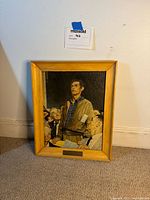 Overall view of framed print leaning against a wall, showing wooden frame and print with plaque visible.