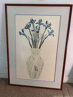 Framed botanical print of blue iris flowers in a vase with thin wood frame leaning against a wall.